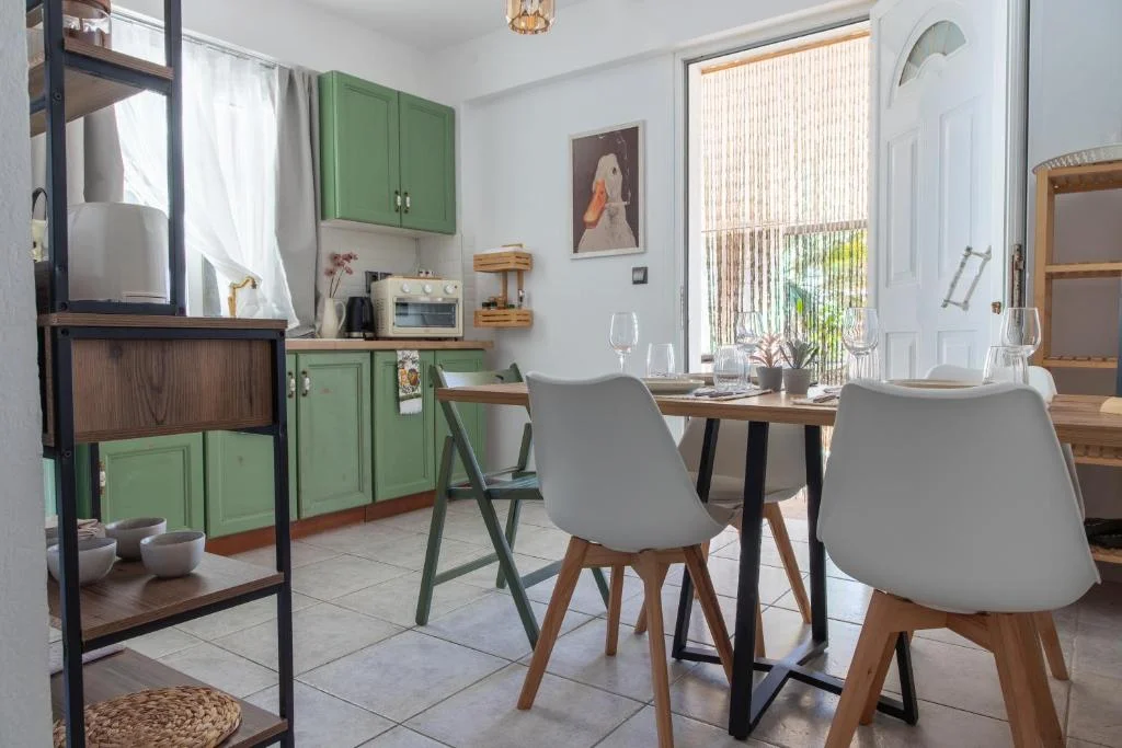 Wooden table with two white chairs and green folding chairs in the kitchen, beside green cabinets and a bright window.