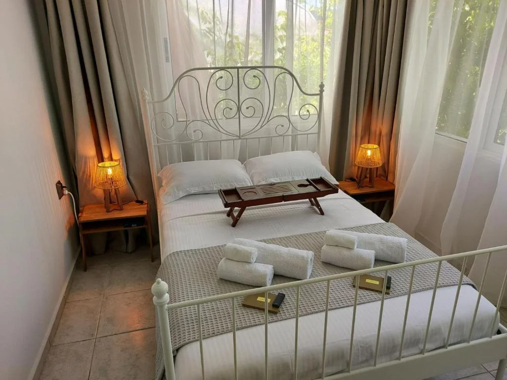 White metal double bed with a wooden tray and rolled towels, between two bedside lamps and curtained windows.