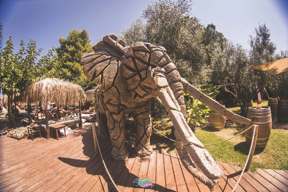 Elephant sculpture beside shaded wooden seating and thatched parasols at Elephant Beach Bar in Pefkochori.